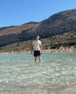 Ein junger Mann in weißem T-Shirt und schwarzen Shorts steht im knietiefen, klaren Wasser vor einer felsigen Berglandschaft.