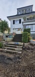 Steinerne Außentreppe führt an einer Baustelle mit Erdaushub hinauf zu einer Terrasse und einem weißen Wohnhaus.