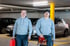 Zwei Handwerker in grauen Hoodies mit Werkzeugkisten in einer Tiefgarage stehen vor zwei Autos.