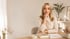 Blonde Frau sitzt lächelnd an einem Schreibtisch mit Notizbuch, Stift und Kaffeetasse in einem hellen, modernen Büro.