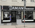 Frontansicht des Damian Barbershops mit Schaufenstern, einem Barbierpfahl und Preistafel für Haarpflege.