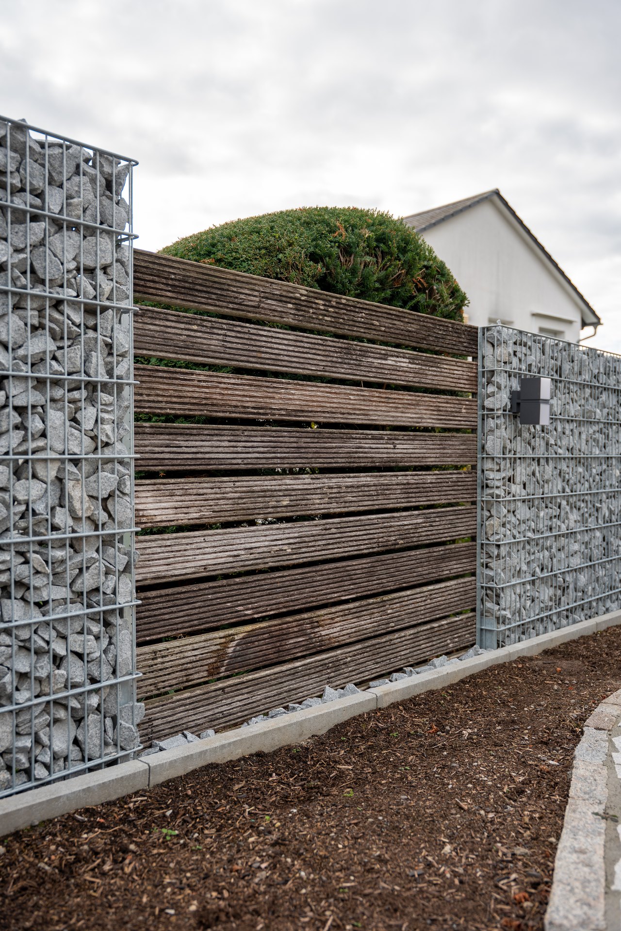 Gartenzaun aus dunklen Holzlamellen zwischen zwei grauen Gabionenwänden mit Steinfüllung vor einem Wohnhaus