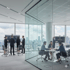 Geschäftsleute in einem modernen Büro mit Glaswänden und Stadtblick, diskutieren vor Bildschirmen und an einem Tisch.