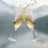 Zwei Sektgläser stoßen an, wobei Sekt herausspritzt, mit verschwommenen Bergen und Wasser im Hintergrund.
