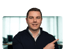 Ein lächelnder Mann in einem dunkelblauen Pullover zeigt mit dem Finger nach rechts.