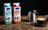 Zwei Milchkartons mit "Frische Fettarme Milch" und "Frische Vollmilch" neben Kaffeetasse und Kaffeebohnen.