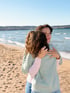 Zwei Frauen umarmen sich am Strand mit einem See und Horizont im Hintergrund.