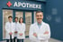 Ein lächelnder Apotheker vor drei Kollegen und dem Schriftzug "APOTHEKE" mit rotem Kreuz auf blauer Fassade.