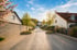 Sonnenbeschienene, gepflasterte Straße zwischen Häusern mit Bäumen und blühenden Sträuchern.
