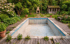 Leerer rechteckiger Swimmingpool mit Rissen, umgeben von Holzsteg, Pflanzen, Gartenhaus und Außenkamin in grünem Garten.