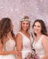 Drei lächelnde Frauen in weißen Spitzen-Brautkleidern vor einem rosa Bokeh-Hintergrund, die mittlere trägt einen Blumenkranz.