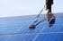 Mann reinigt Solarpanel mit langem Schrubber und Wasser unter blauem Himmel.
