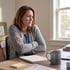 A woman with a furrowed brow sits at a desk with crossed arms, looking out a window next to a mug and notebook.