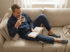Ein lächelnder Mann in blau kariertem Pyjama liest ein Buch auf einem beigen Sofa und trinkt aus einer Tasse.