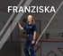 FRANZISKA. Frau in blauer Arbeitshose und T-Shirt mit Logo, steht in einem Büro und lächelt.