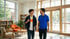 A smiling student with a backpack and teacher in a blue polo shirt walk through a bright school library or lounge.