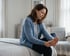 A woman in a blue cardigan sits on a bed and rubs her bare foot with both hands while looking down with a somber expression.