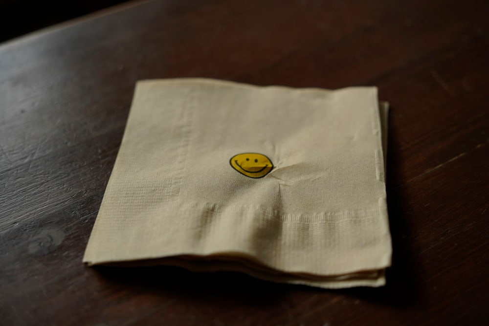 Eine zerknitterte Papierserviette mit einem kleinen gedruckten Smiley liegt auf einem dunklen Holztisch.