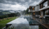 Modernes Haus mit Infinity-Pool unter bewölktem Himmel, umgeben von Wald und Bergen.