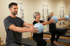 Drei Personen machen Pilates mit blauen Bällen in einem Fitnessstudio mit Reformern im Hintergrund.