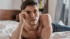 Man with dark hair and bare chest lying on a white bed, looking forward.