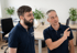 Zwei Männer in dunkelblauen Poloshirts mit "PFLEGE LEICHT" Logo unterhalten sich in einem Büro. Der eine zeigt nach rechts.