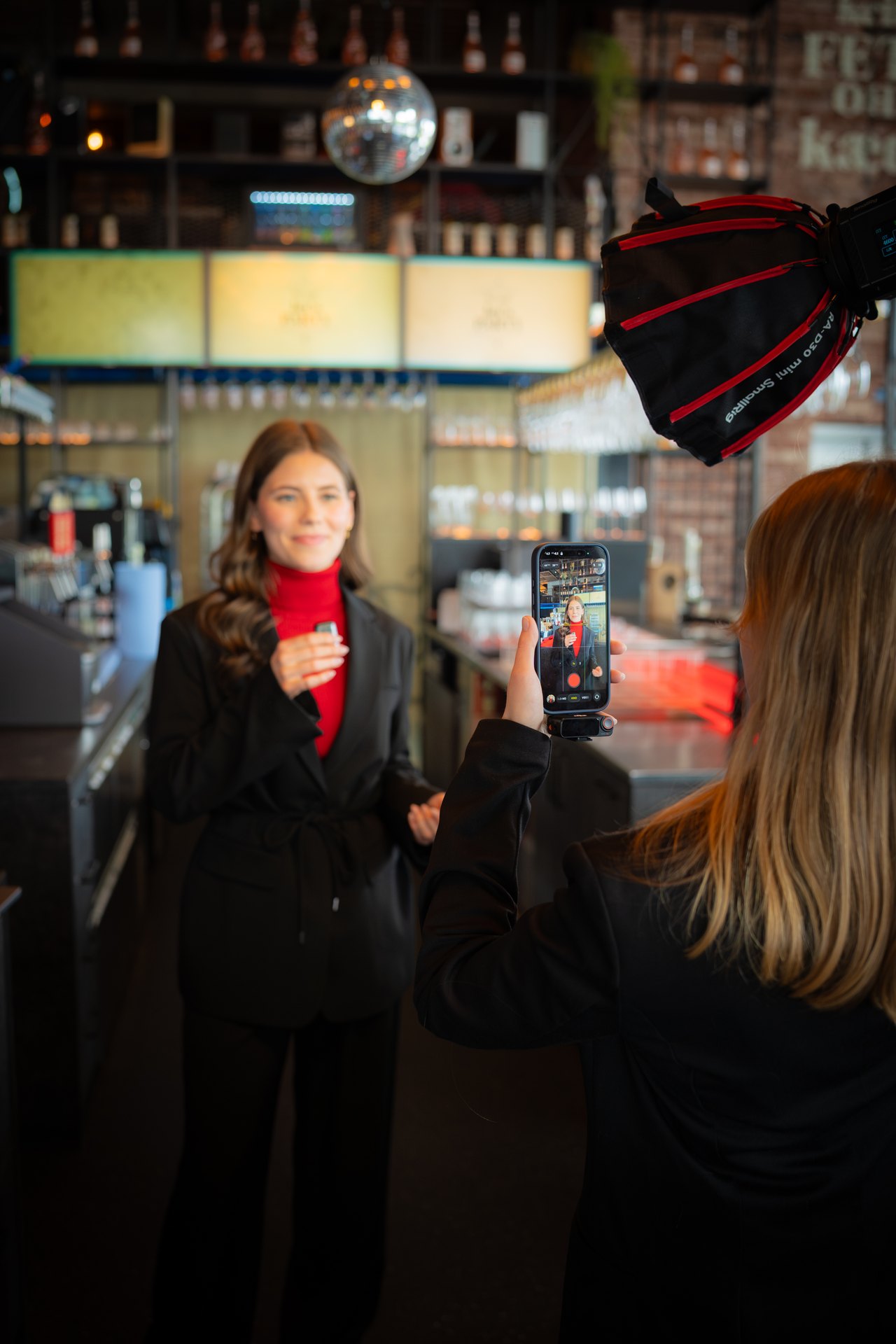 Eine Frau filmt eine andere Frau in einem roten Rollkragenpullover und einem schwarzen Blazer mit einem Smartphone.