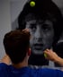 Person wirft Tennisbälle auf ein schwarz-weißes Poster von Sylvester Stallone als Rocky.