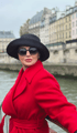 Frau im roten Mantel mit schwarzem Hut und Sonnenbrille vor einem Fluss und Pariser Stadthäusern.