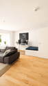 Modernes Wohnzimmer mit Parkett, dunklem Sofa, weißem Schreibtisch mit zwei Monitoren und einem ausziehbaren Tagesbett.