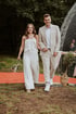 A smiling couple walks hand-in-hand down a red carpet outdoors, woman in white jumpsuit, man in beige suit.
