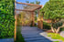 Holzpergola und Steinpfad in einem Garten mit grünen Pflanzen, Moos und einem Baum.