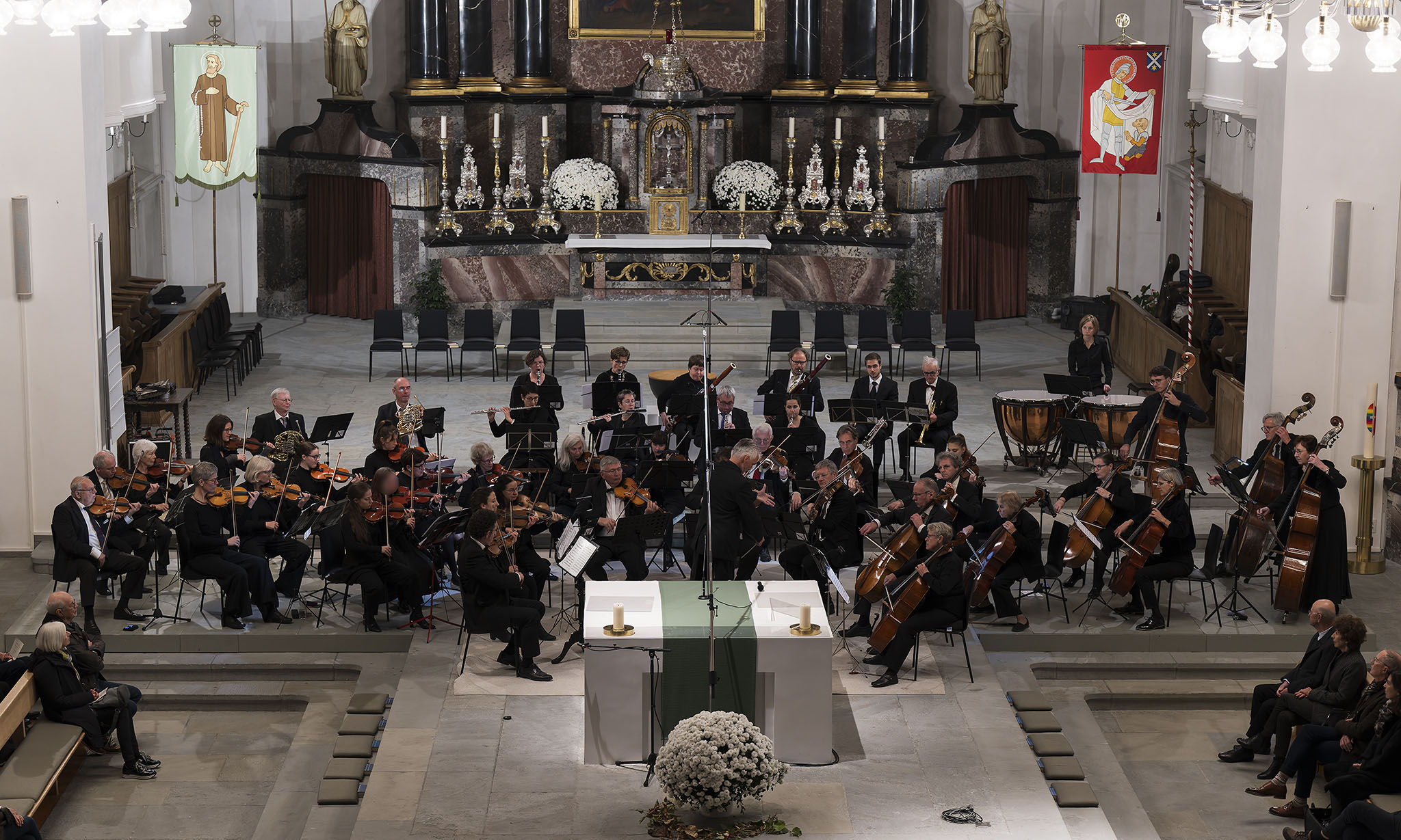 Ein klassisches Orchester spielt ein Konzert in einer hellen Kirche vor einem prunkvollen Altar und Publikum.