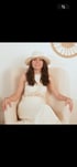 Woman with long dark hair, wearing a white straw hat and a white bohemian-style outfit, sitting in an armchair.