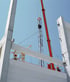 Construction workers in hard hats guide a large concrete beam being lowered by a crane at a building site under a blue sky.