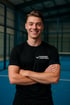 Lächelnder junger Mann mit verschränkten Armen und einem schwarzen T-Shirt mit der Aufschrift "PADEL FACTORIA" auf einem blauen Padel-Platz.