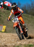 Ein Motocrossfahrer in orangefarbener Kleidung und Helm fährt ein Dirtbike mit der Nummer 98 auf einer staubigen Strecke.