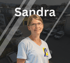 Sandra in hellem T-Shirt mit Logo vor unscharfem Hintergrund eines Gebäudes und Parkplatzes.