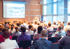Menschen sitzen in einem Konferenzsaal und schauen auf eine Leinwand mit einer Präsentation und einen Redner.
