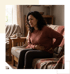 A woman sits on a sofa holding her stomach and lower back with a grimacing facial expression.