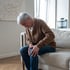 Elderly man in brown cardigan sitting on sofa, holding knee in pain from autoimmune disease