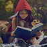 A young girl in a red hooded cape reads a book, with a teddy bear and monkey plush toy beside her.