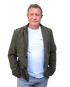 Ein Mann mit grauen Haaren, khakifarbenem Blazer, weißem T-Shirt und Jeans steht mit den Händen in den Taschen.