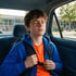 A boy in a blue hoodie and orange shirt sits in a car with his eyes closed, wearing a backpack in front of a school.