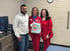 Three healthcare professionals smile while holding a "What Fun" gift bag in a clinical room with dumbbells and a brick wall.