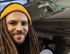 Mann mit Dreadlocks und gelber Mütze lächelt vor einem schwarzen Auto.