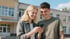 Zwei lächelnde Teenager schauen auf ein Smartphone vor einem Schulgebäude.