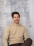 Ein lächelnder junger Mann in einem beigefarbenen Kapuzenpullover sitzt vor einer grau-blauen Wand.