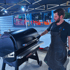 Mann mit Bart und Tätowierungen brät Fleischstücke auf einem großen runden Grill.
