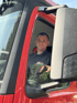Ein Mann im roten LKW mit schwarzem Poloshirt und Firmenlogo lächelt in die Kamera.